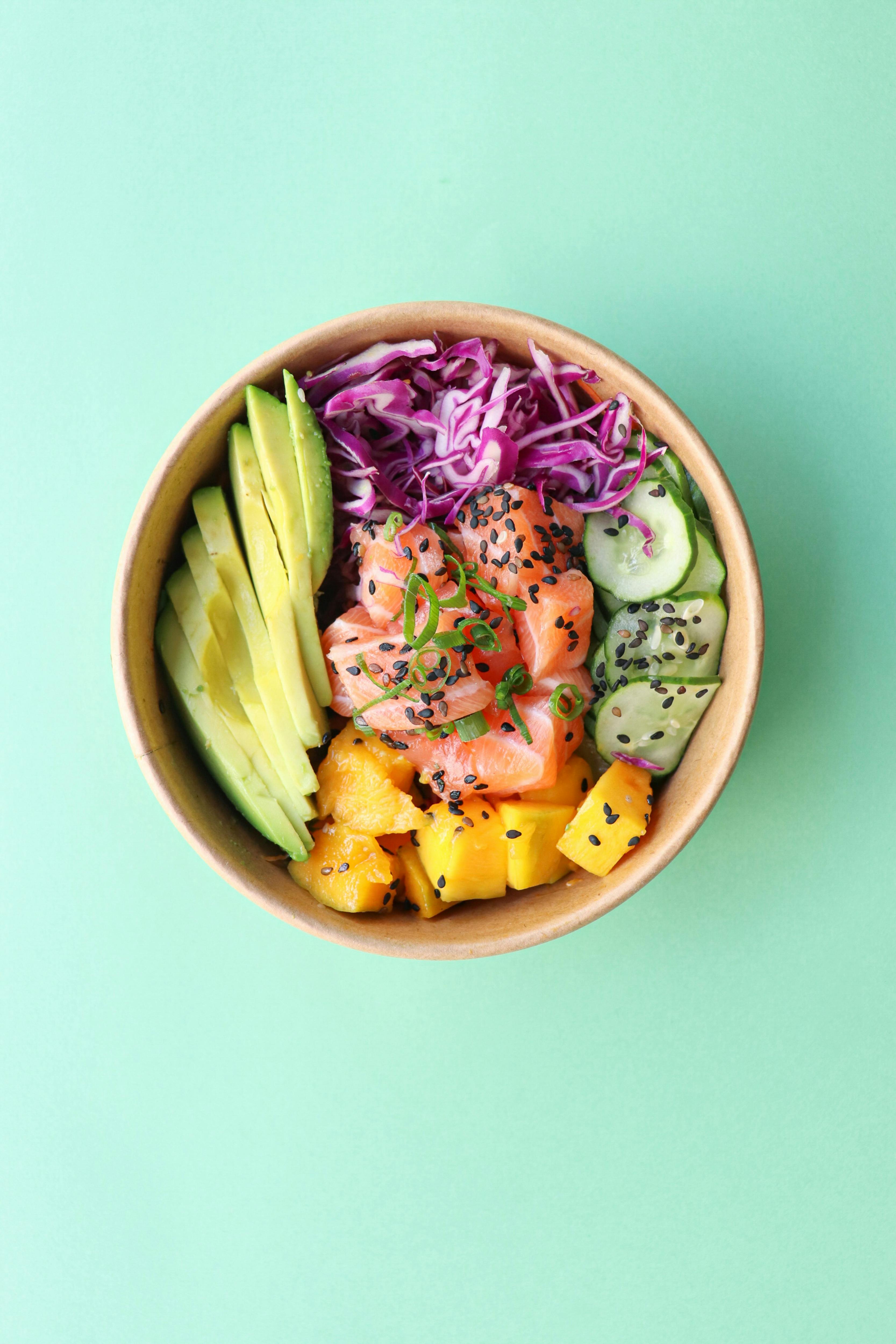 Bowl with sliced avocado, purple cabbage, diced mango, cucumber slices, and raw salmon topped with black sesame seeds and green onions on a light green background.