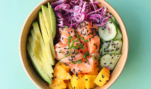 Bowl with sliced avocado, purple cabbage, diced mango, cucumber slices, and raw salmon topped with black sesame seeds and green onions on a light green background.