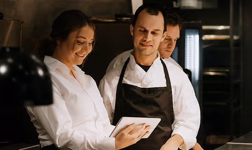 Three chefs in white uniforms and black aprons looking at a tablet and smiling in a professional kitchen.