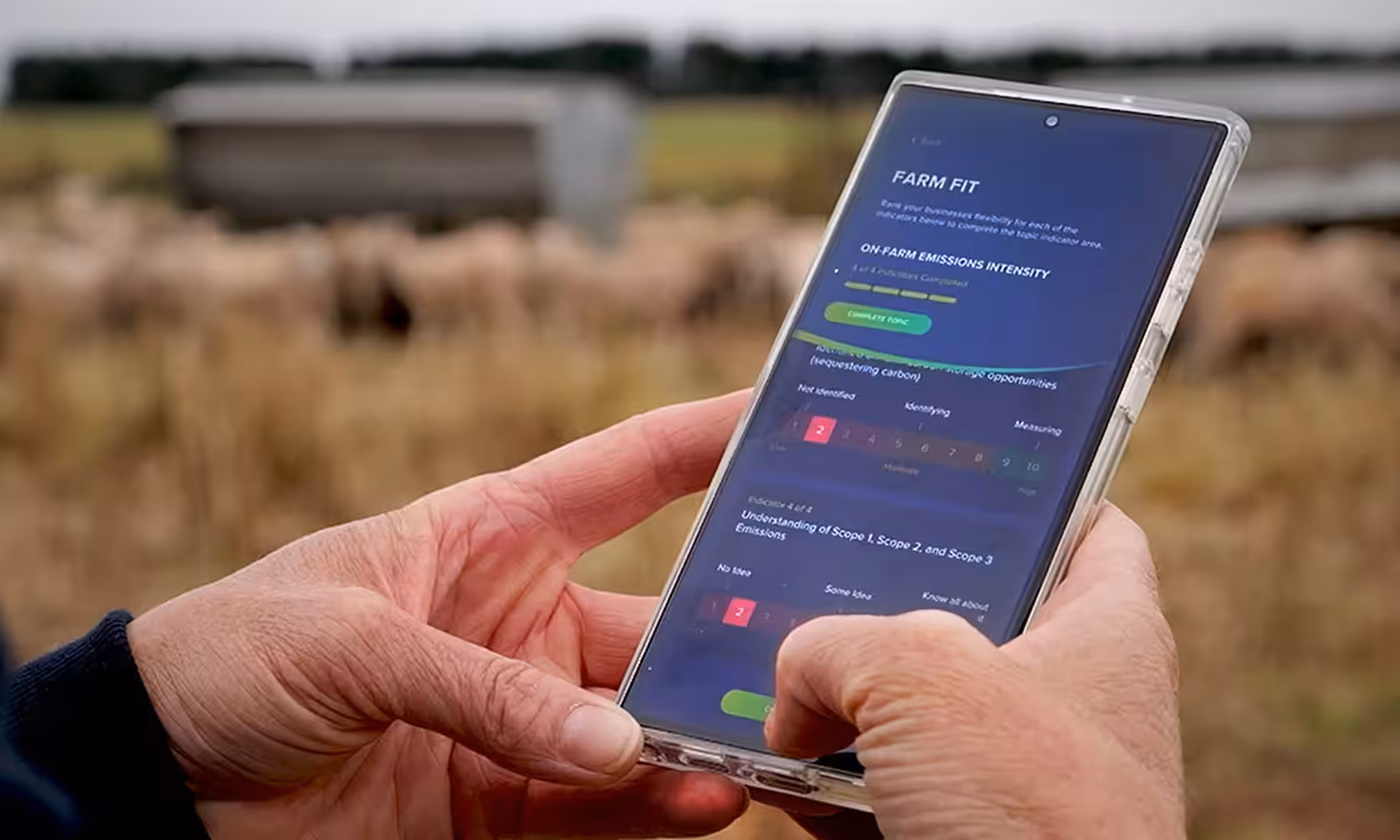 Close-up of hands holding a smartphone displaying the Farm Fit app, showing an on-farm emissions intensity assessment with sliding scale ratings, set against a blurred background of sheep grazing in a paddock.