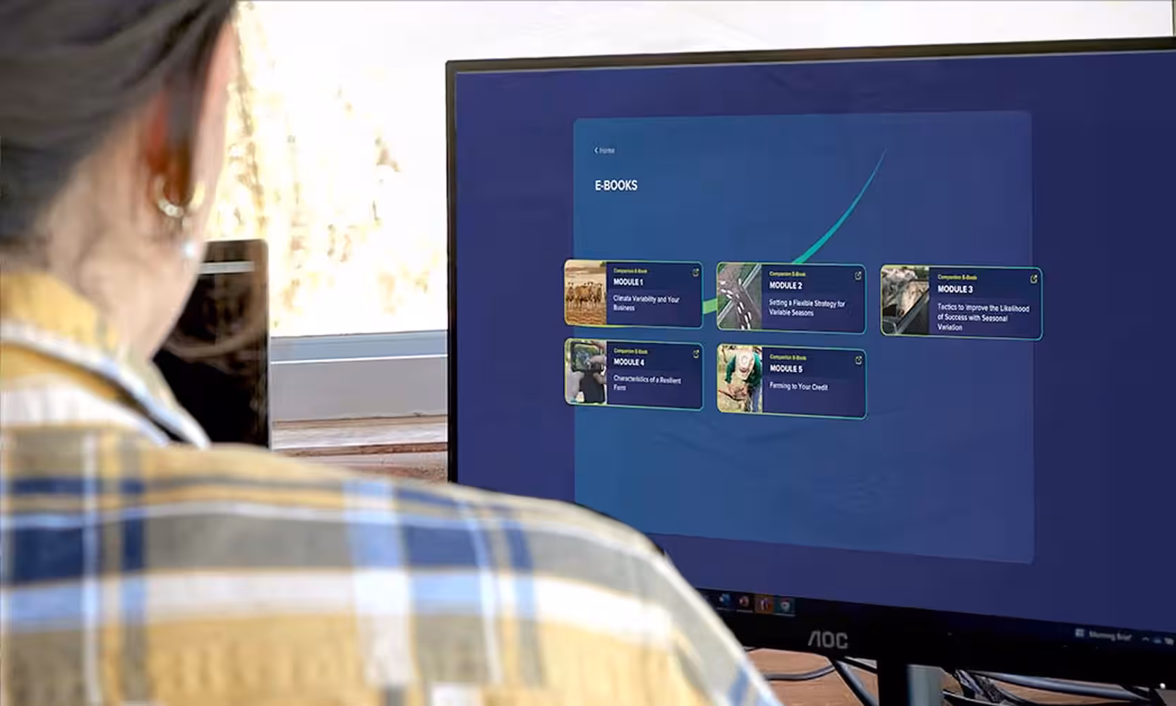 Person viewing the E-Books module selection screen on a desktop monitor, showing five companion e-book modules covering climate variability, flexible strategy, seasonal variation tactics, resilient farming, and farming to your credit.