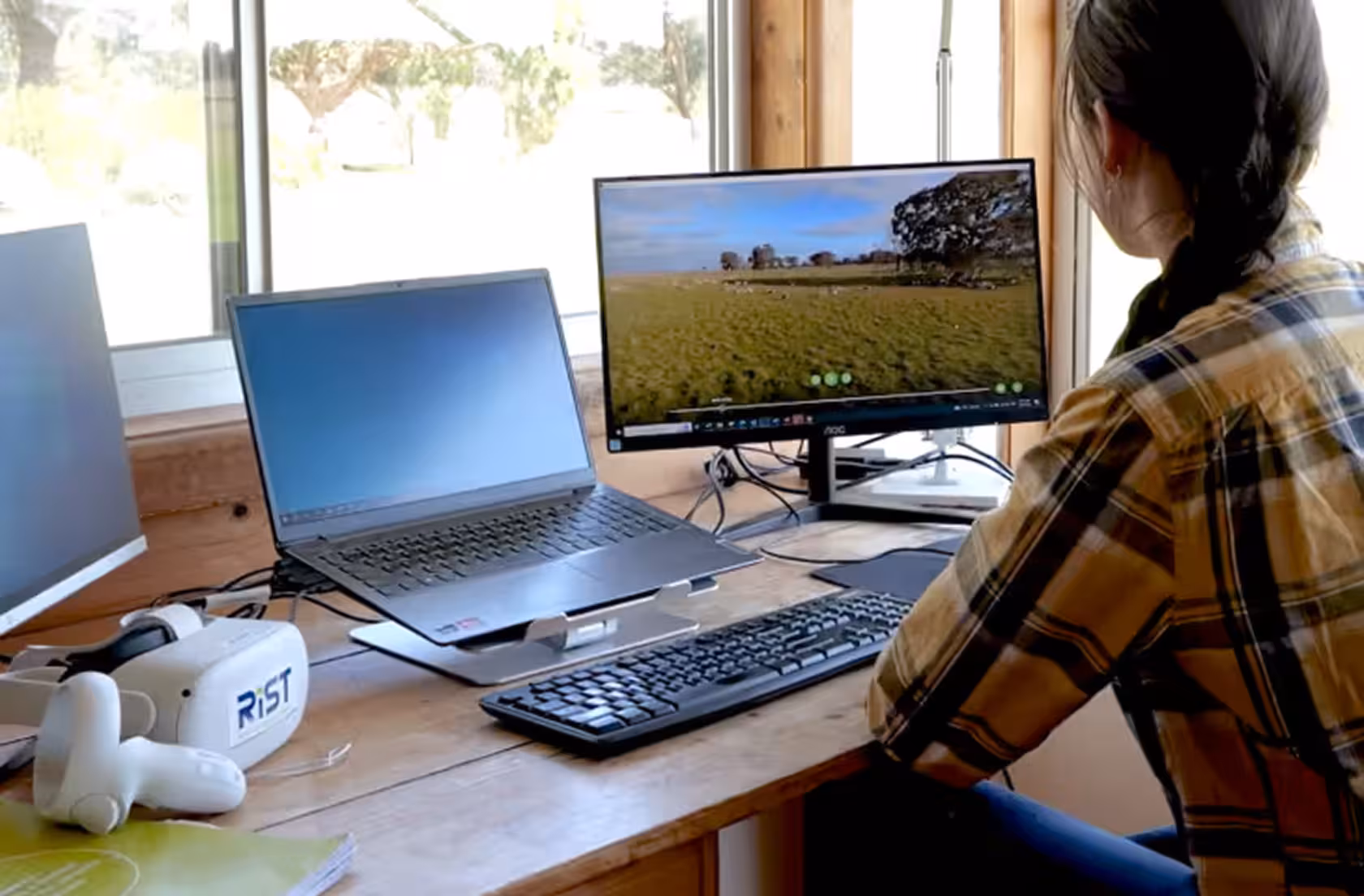 Person seated at a desk viewing a 360° farm paddock scene on a monitor, with a laptop, keyboard, and RIST-branded VR headset and controllers on the desk beside them.