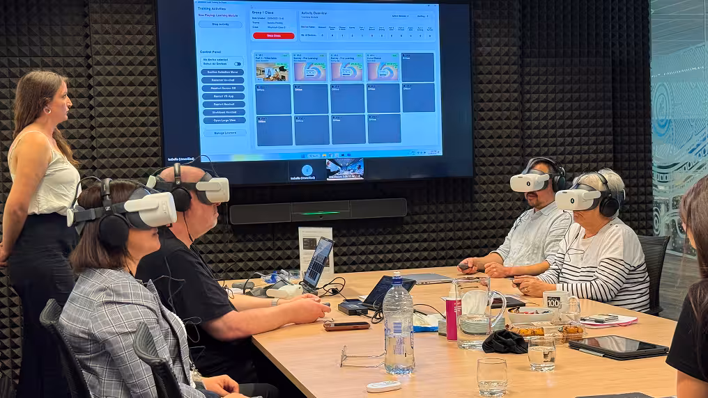 A group of people seated at a conference table wearing VR headsets with headphones during a training session, while a facilitator stands and speaks beside a large screen displaying a training activity dashboard with device status and live video feed from learners' app.