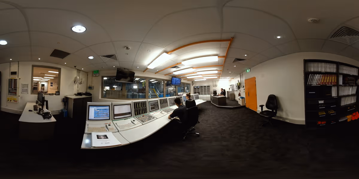 360-degree panoramic view of a nuclear facility control room with a long curved desk of computer monitors and control panels, two staff seated at the desk, and a reception area with a third person visible in the background.