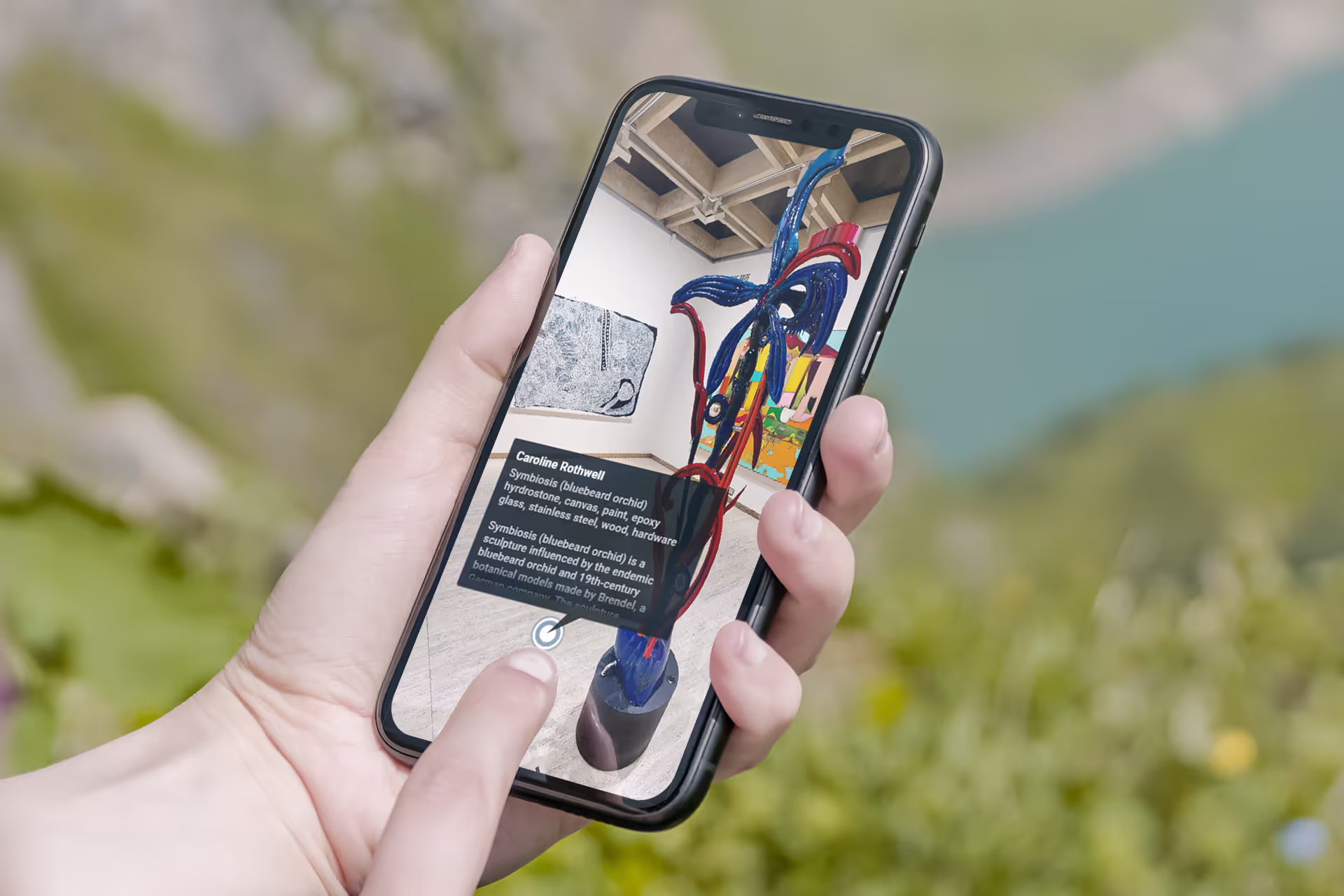 A person holds a phone displaying a Matterport tour with an info panel for Caroline Rothwell's sculpture "Symbiosis (bluebeard orchid)."