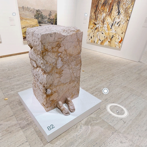 Matterport tour view of a stone block sculpture with carved feet, displayed on a white plinth in the gallery.