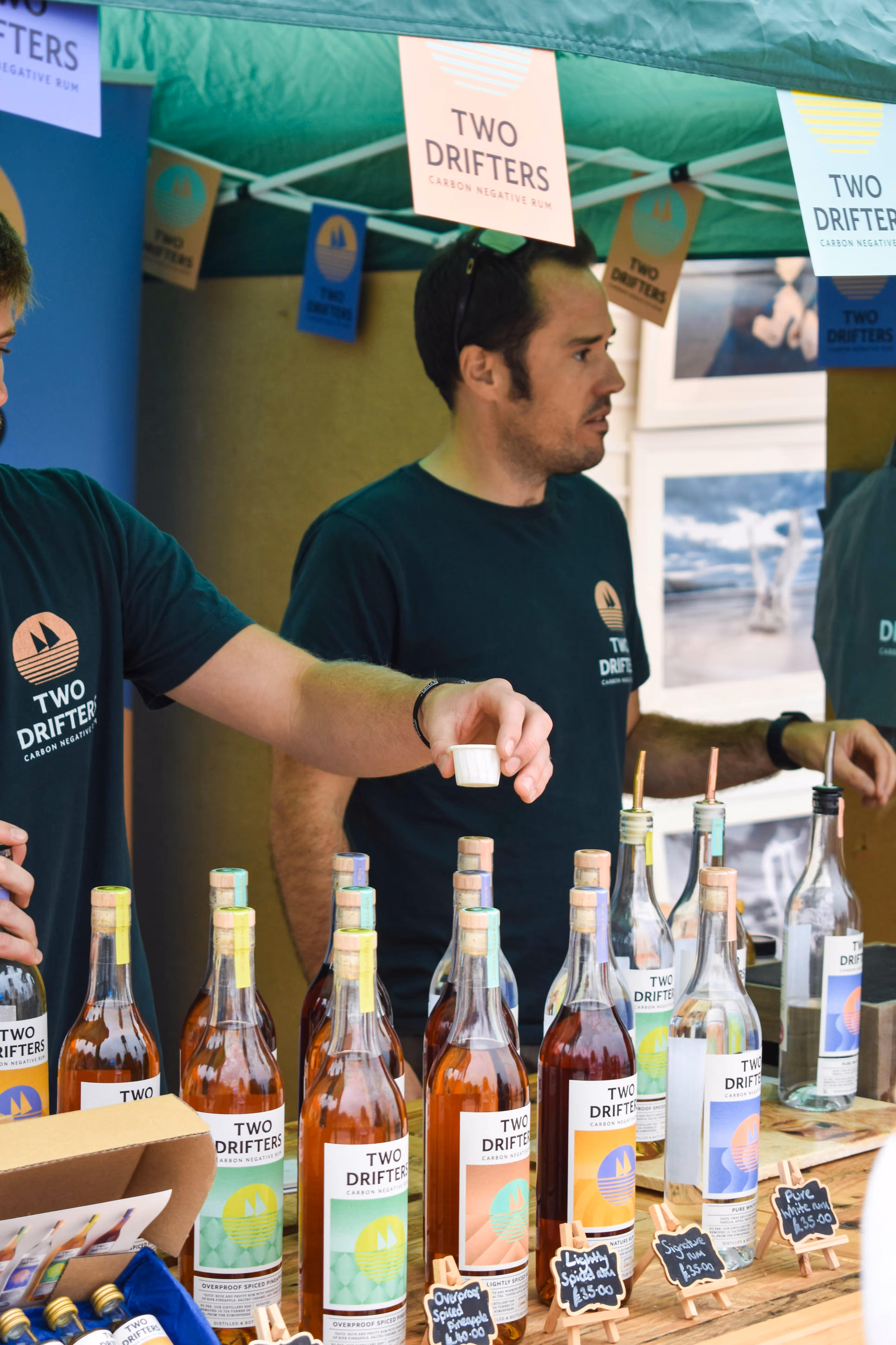 Man in Two Drifters shirt holding a small cup at a rum tasting stall with bottles and signs.