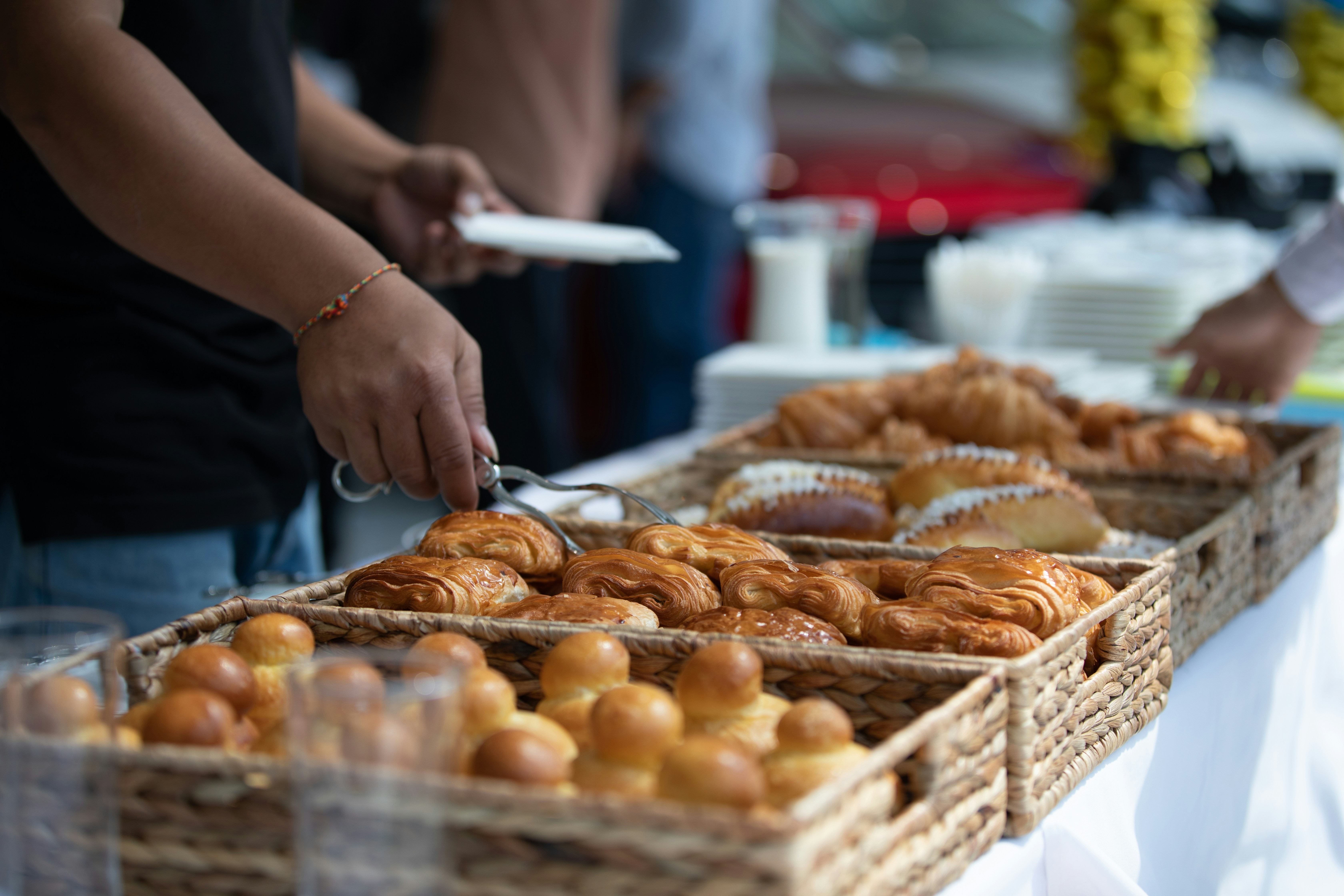 Hand using tongs to pick pastry from woven baskets filled with assorted breads on a white tablecloth.