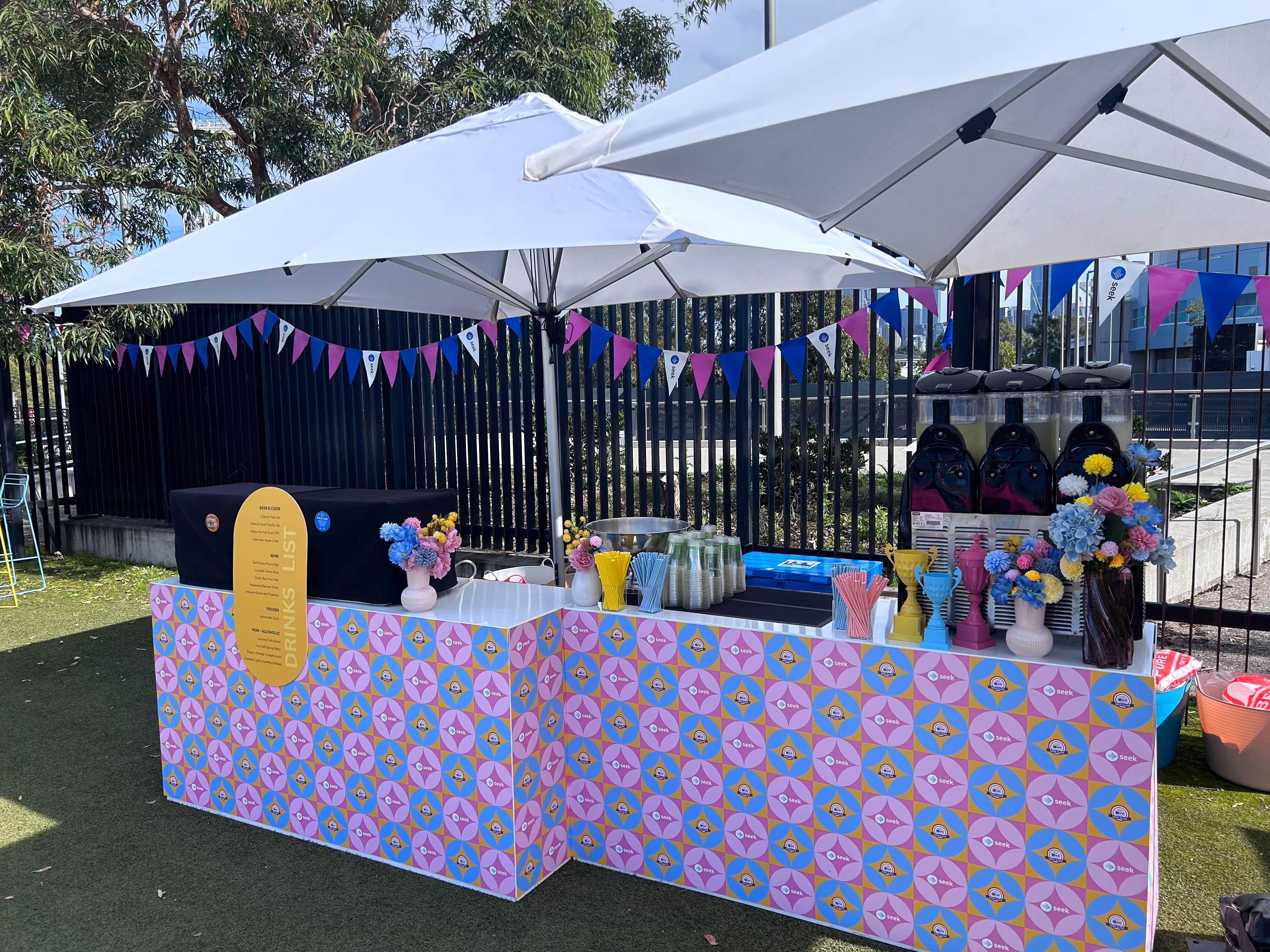 Outdoor drinks stall with patterned counters, shaded by large white umbrellas and decorated with flowers and flags.