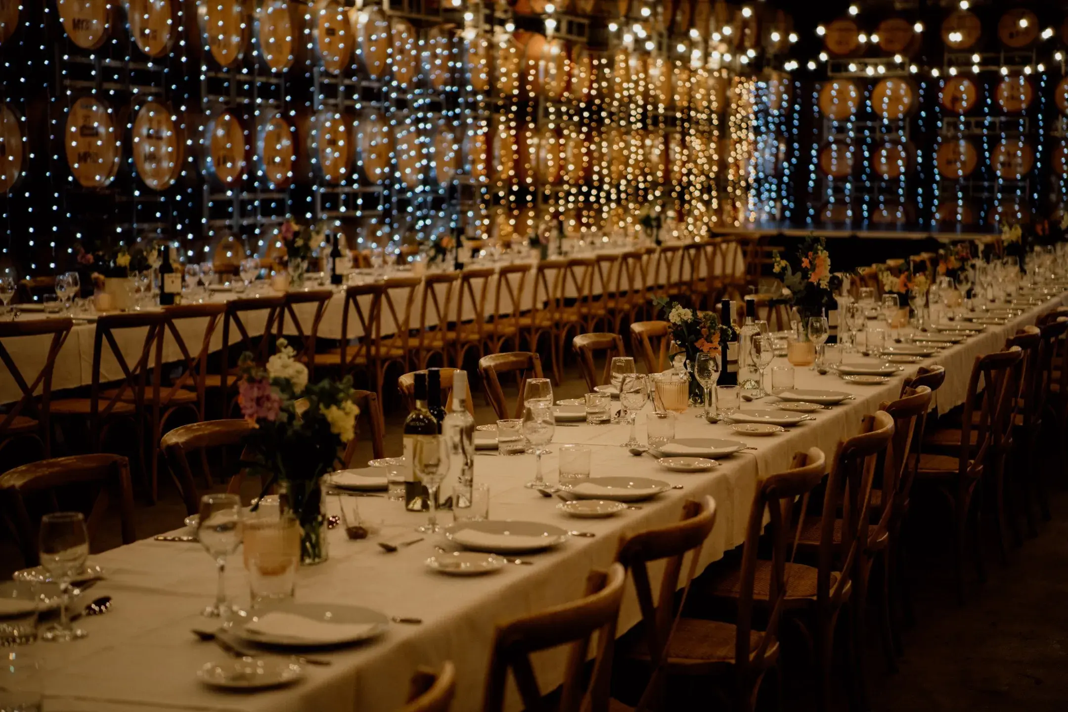 Long banquet tables set with white linens, plates, glassware, flowers, bottles, and string lights in background.