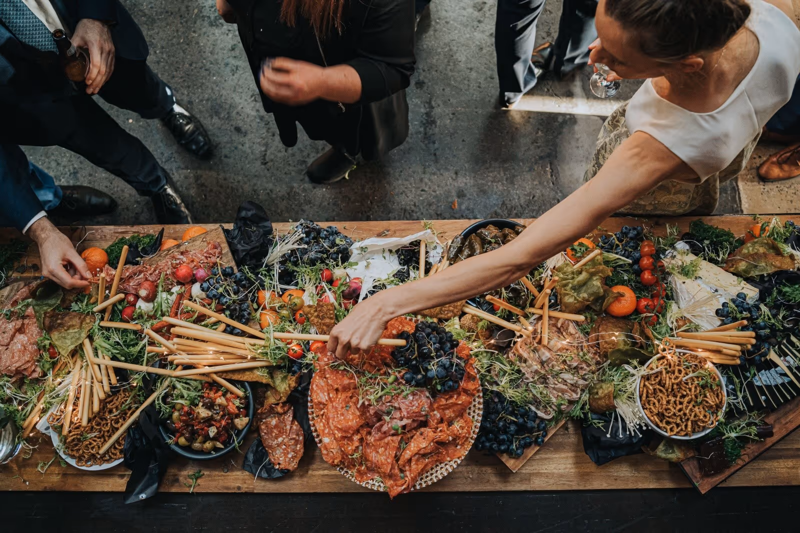 Overhead view of people reaching for snacks on a large charcuterie board with meats, grapes, and breadsticks.