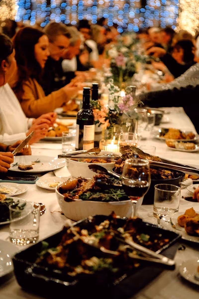 People seated at a long table enjoying a meal with wine, candles, and floral centerpieces.
