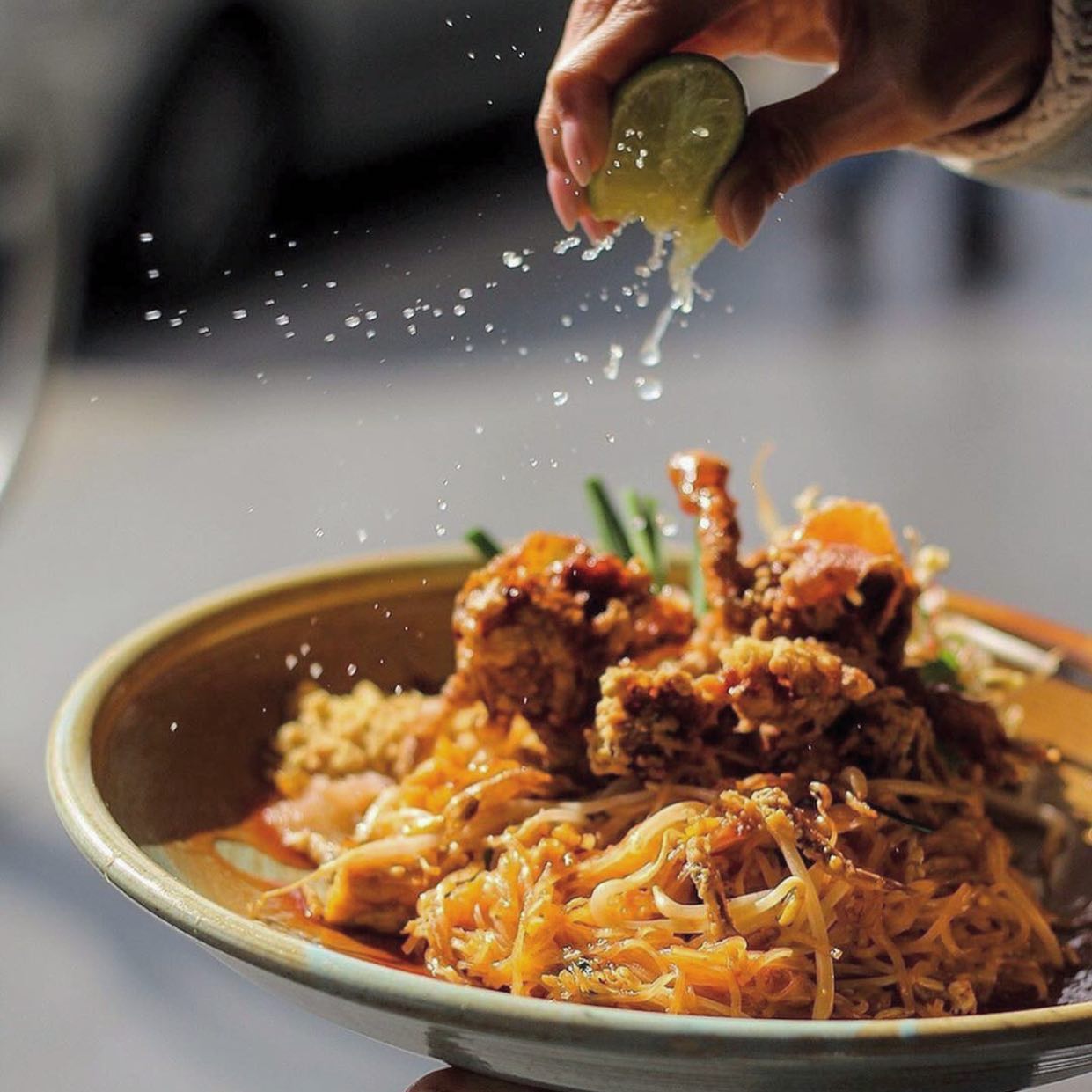 Hand squeezing lime over a bowl of noodles topped with fried chicken and greens.