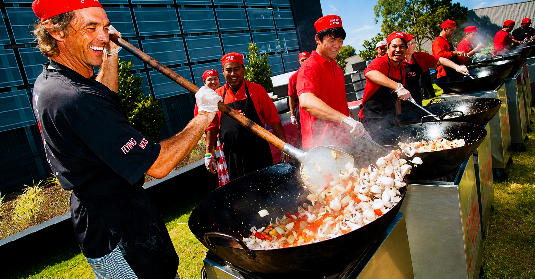 Smiling chefs wearing red uniforms cooking vegetables in large woks outdoors in a row on a sunny day.