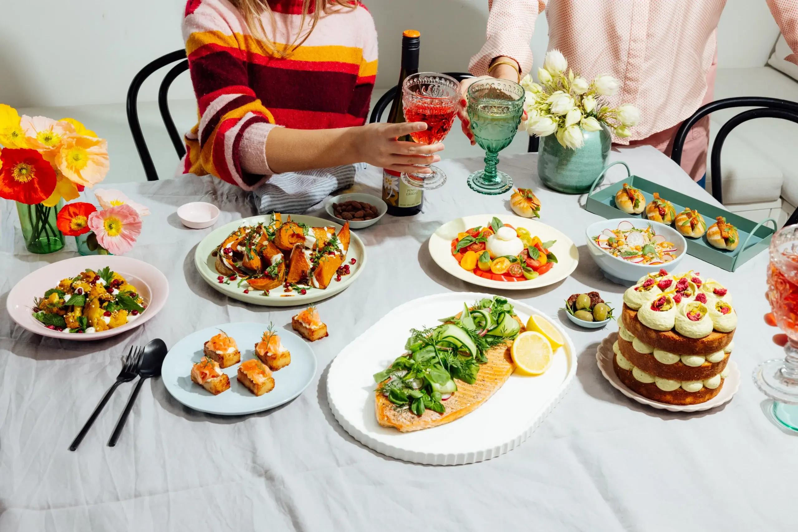 Two people clinking glasses over a table set with diverse colorful dishes and flowers.