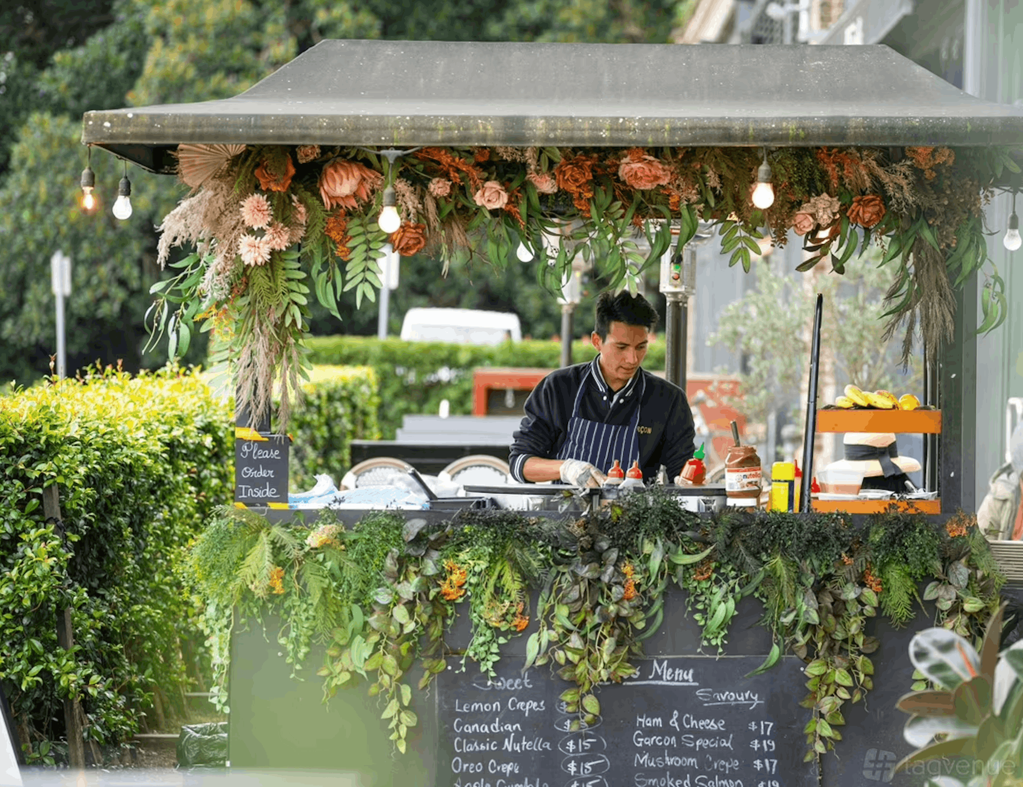 Man wearing apron preparing food at a flower-decorated outdoor food stall with menu board.