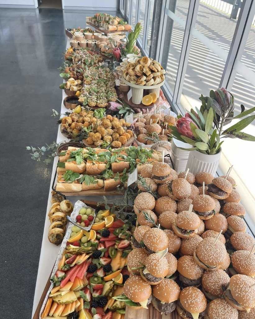 Long table with assorted sliders, sandwiches, fruit platter, pastries, and appetizers near large windows.