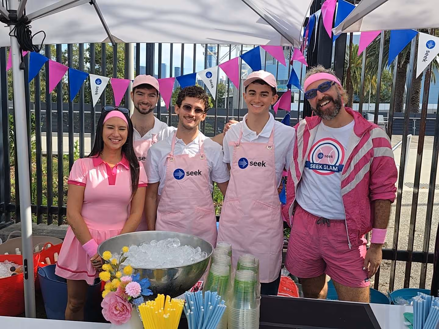 Five people wearing pink Seek aprons and outfits smile behind a table with ice and straws outdoors.
