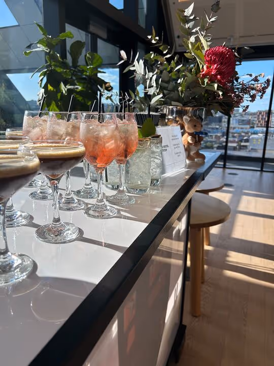 Row of assorted cocktails on a bar counter with plants and large windows in the background.