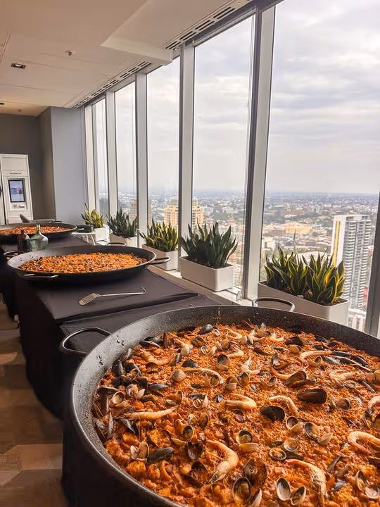 Three large pans of seafood paella on a table by tall windows overlooking a cityscape.