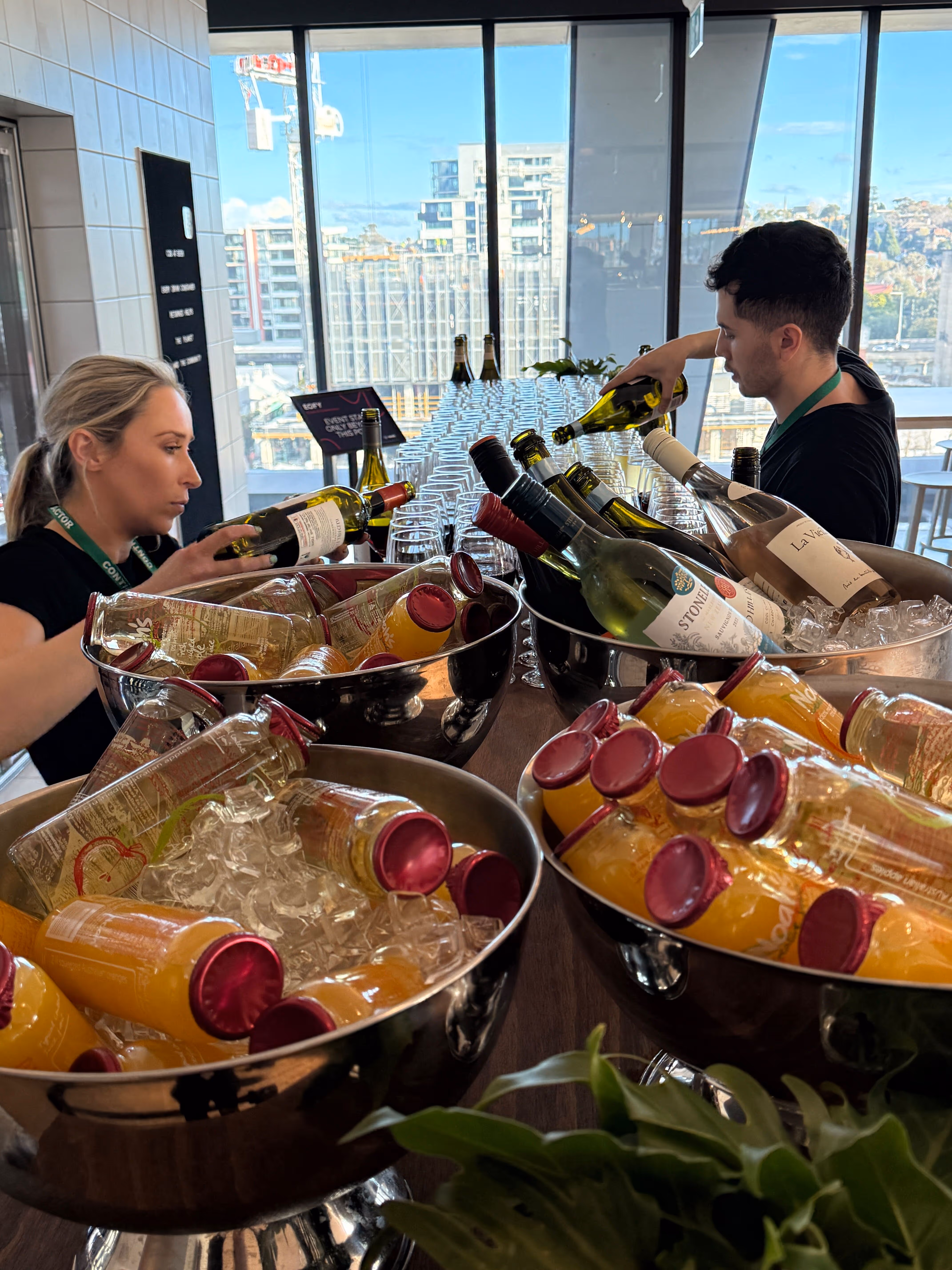 Two servers pouring white wine behind a table with glasses and buckets of chilled bottles and juice.