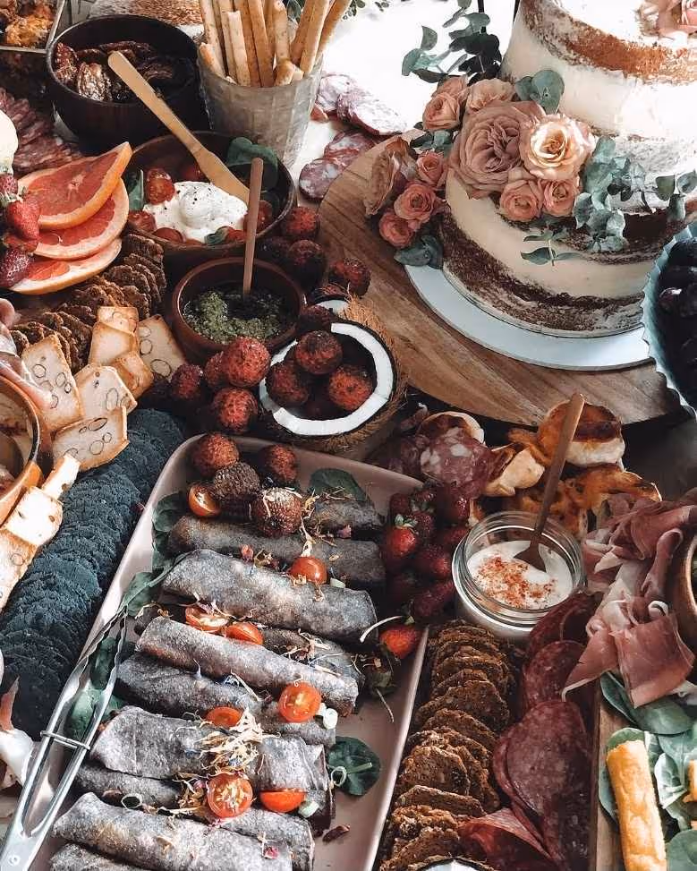 Rustic wedding charcuterie spread with cake, crackers, fruits, dips, and savory rolls on wooden board.