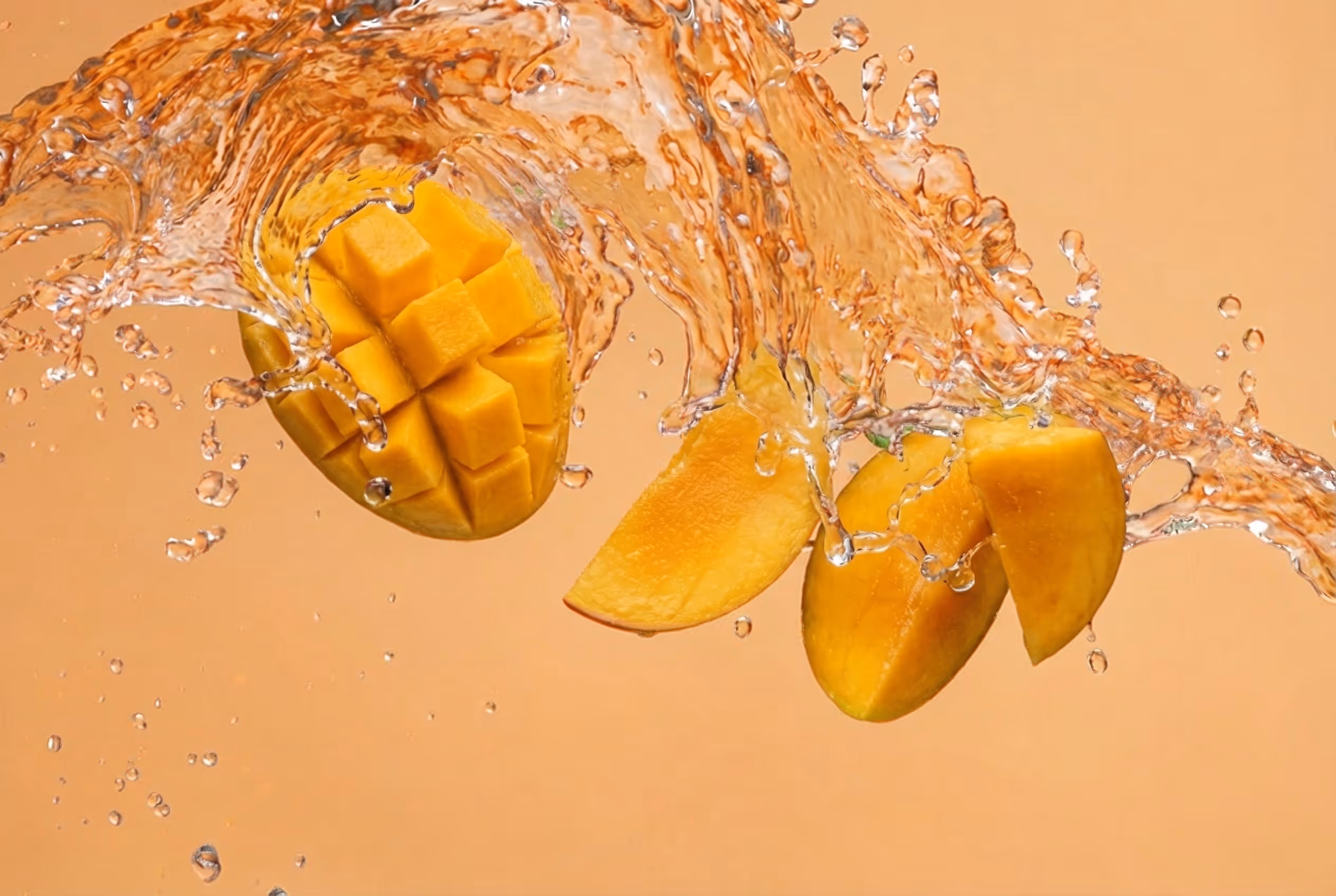 Cubed and sliced mango pieces splashing in clear water against a peach background.