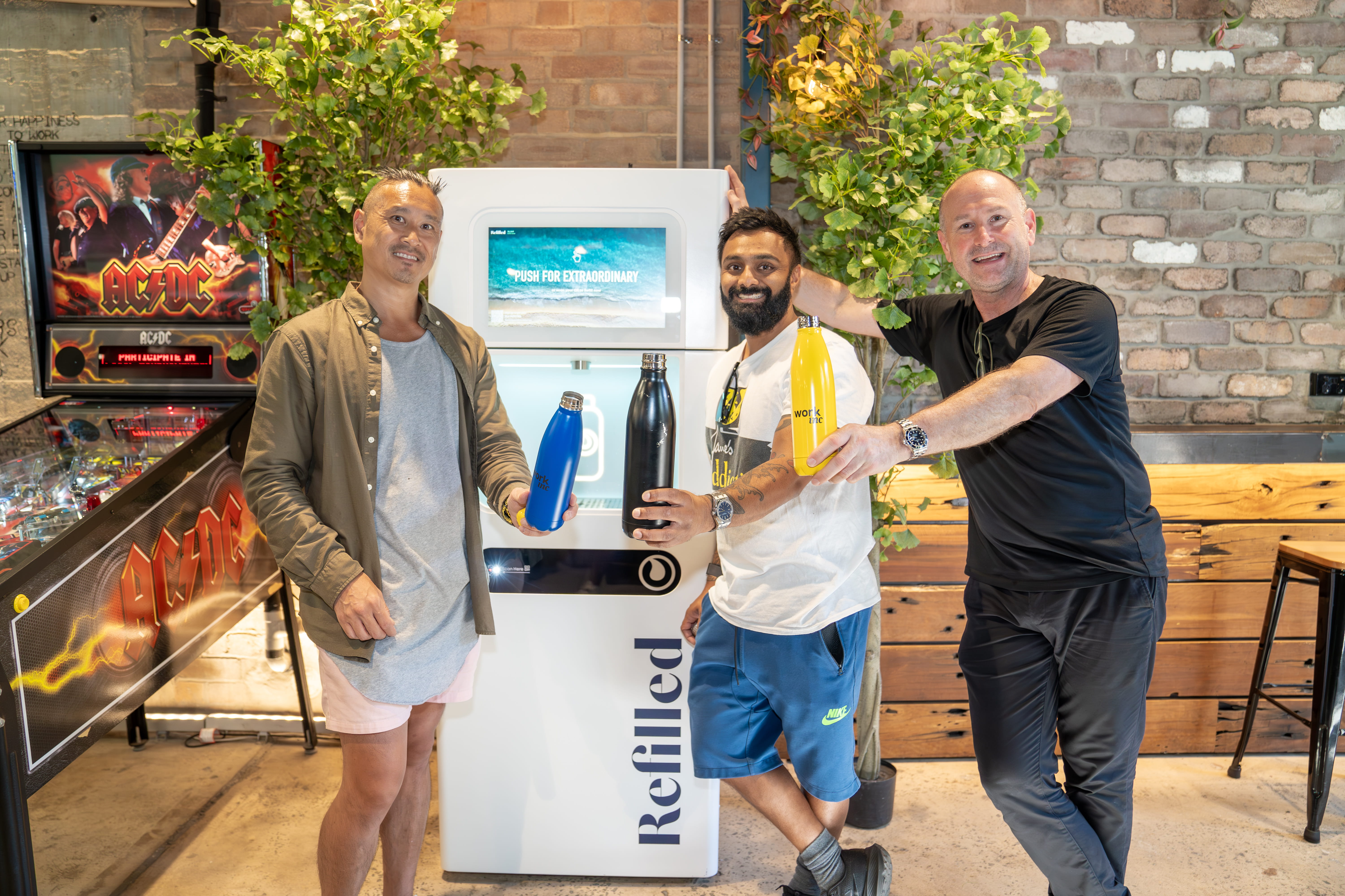Three men smiling, holding reusable bottles in front of a Refilled bottle-filling station and greenery.