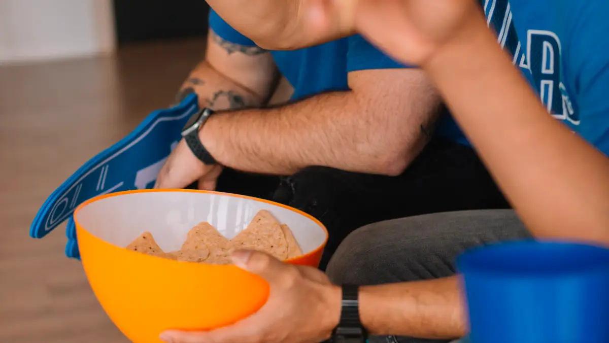 Football fans watching the game wearing blue and eating chips from an orange bowl.