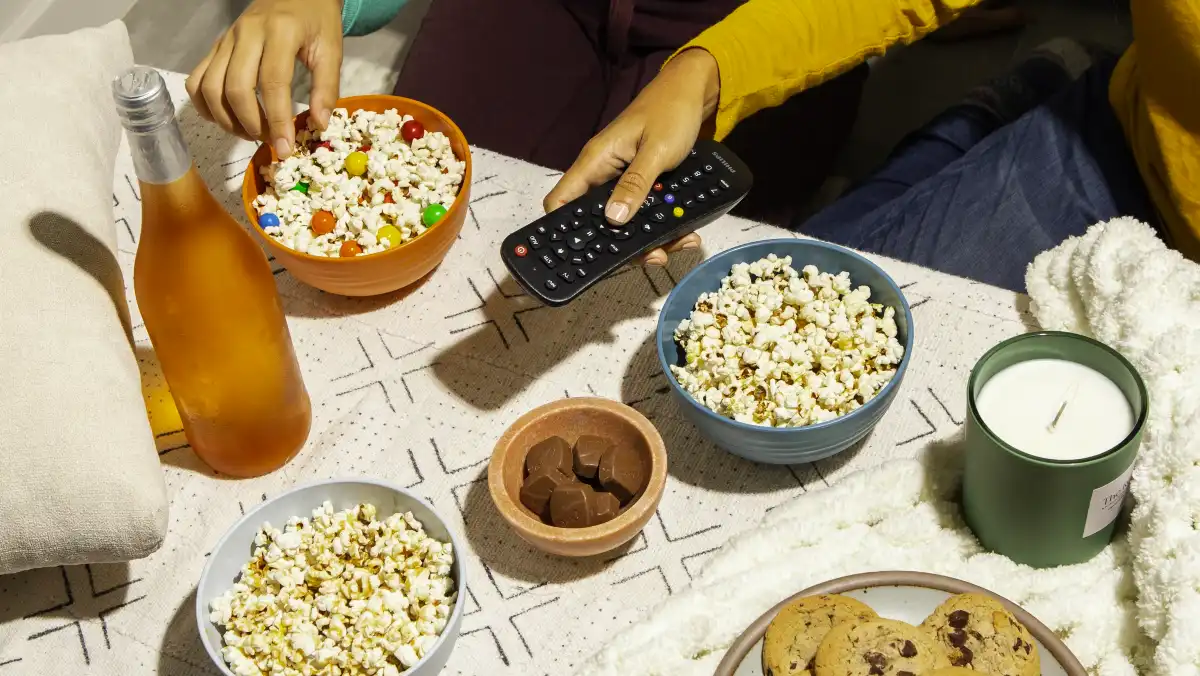 A table full of snacks including popcorn, chocolate and cookies and hands holding a remote control.
