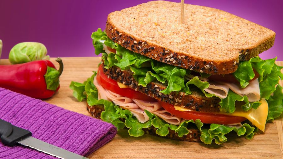 A sandwich with tomato, lettuce, ham and cheese sitting on a wooden block with a purple background.