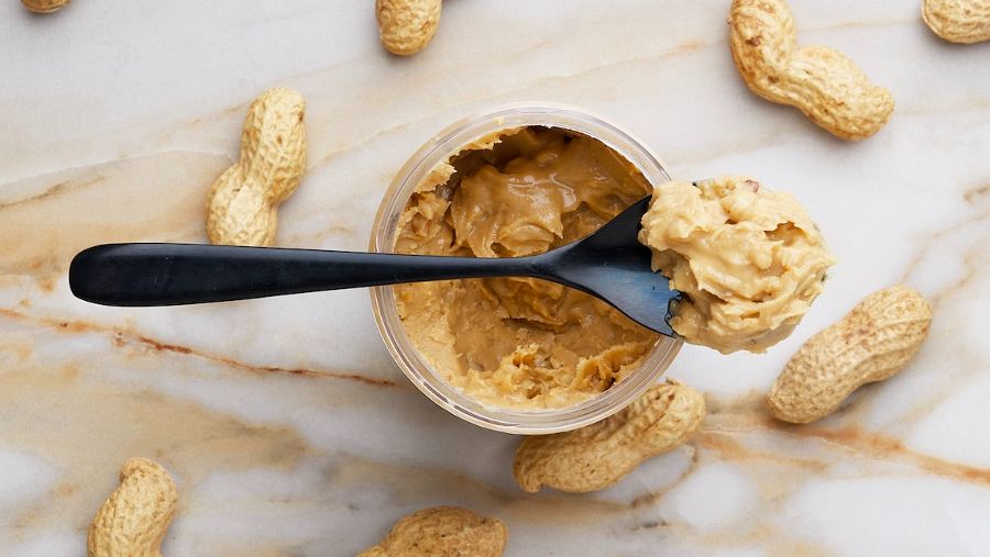 A peanut butter jar with a spoon resting across it with a scoop of peanut butter on it. It's resting on a marble table with peanuts on it still in the shell.