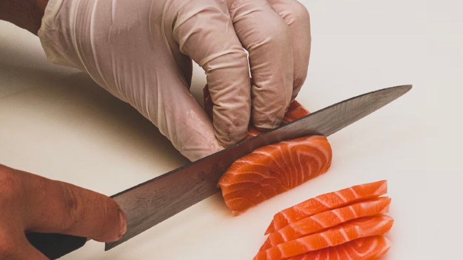 A gloved hand cutting up salmon into sashimi.