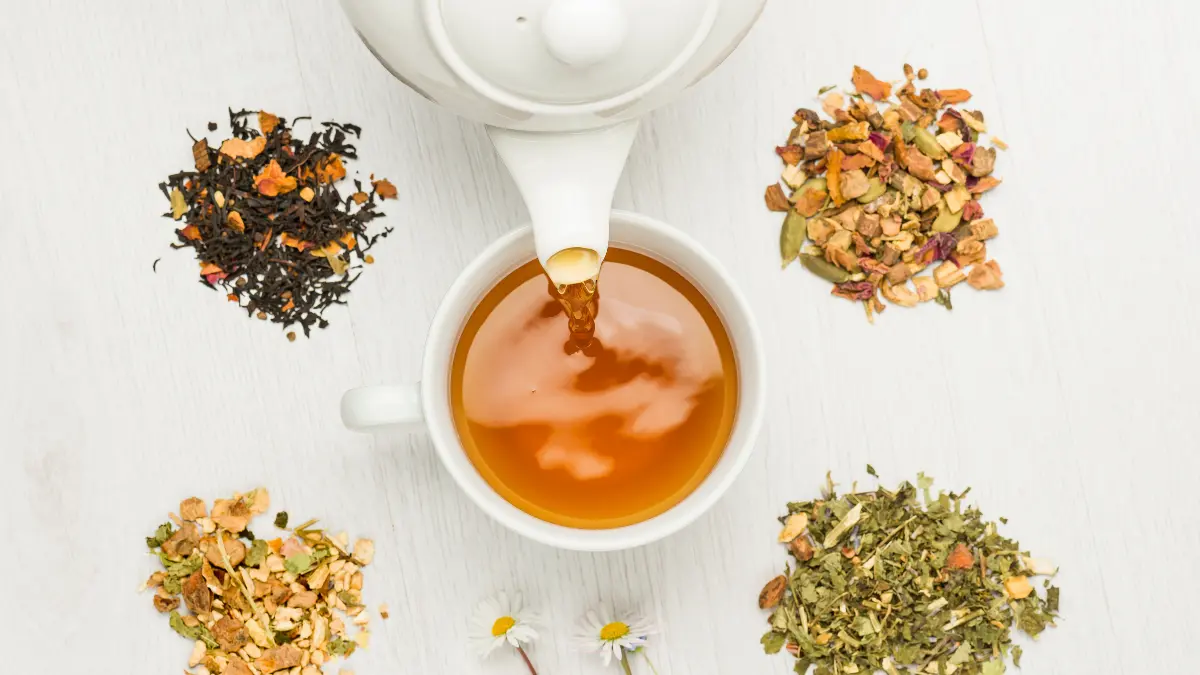 A tea pot pouring tea into a cup that's surrounded by various loose tea leaves.