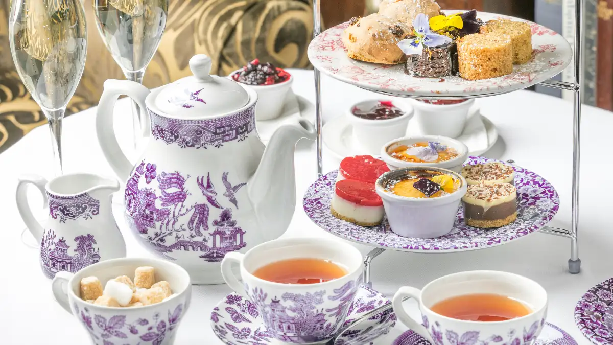 An elegant teapot with two cups of tea next to a tiered high tea stand with various sweet treats.