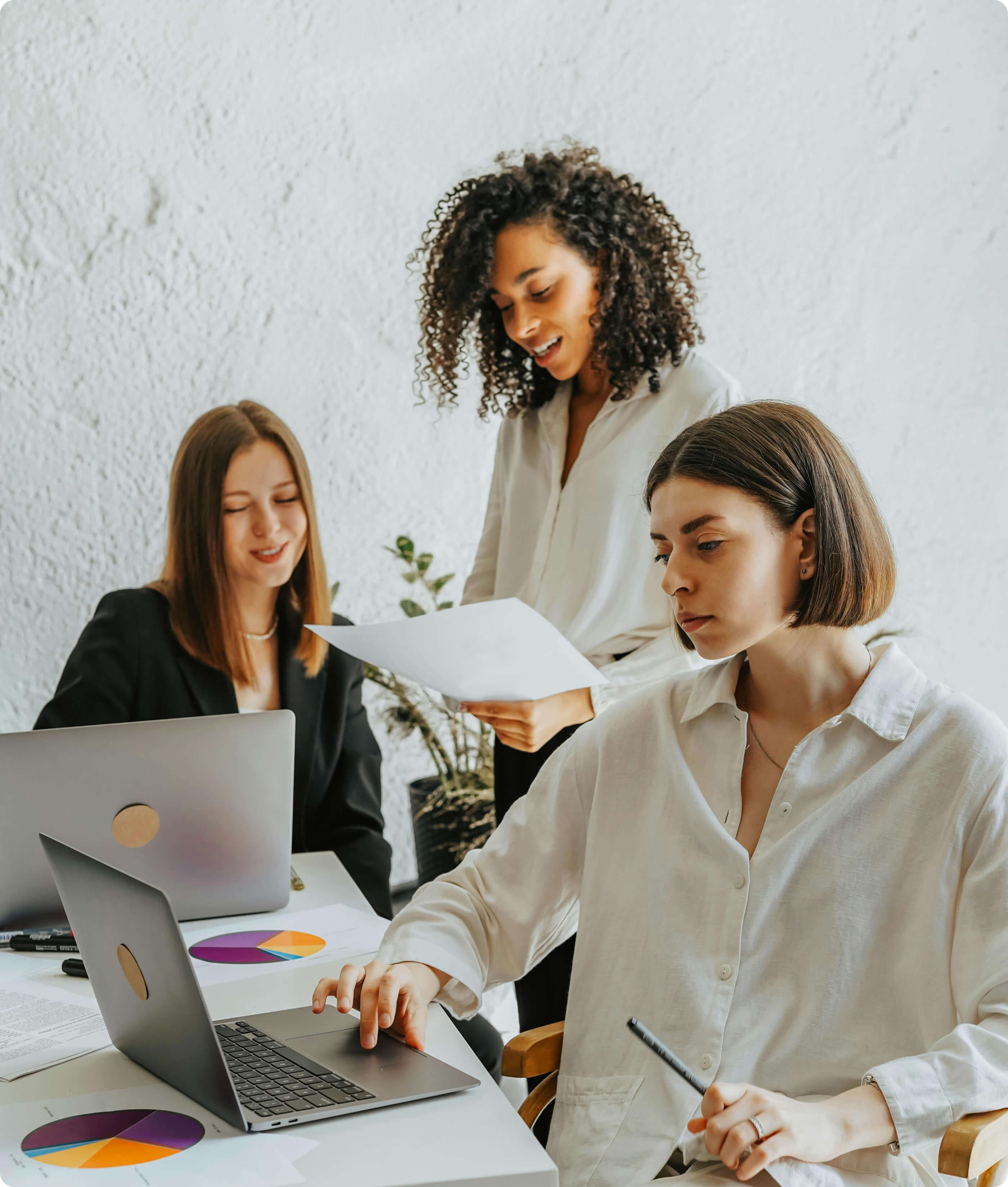 Three women collaborating in a bright office, reviewing documents and working on laptops with colorful pie charts on the table.