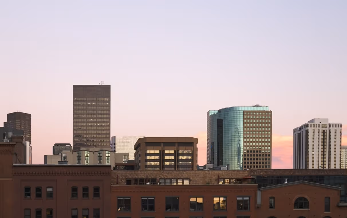 City skyline with modern glass and brick buildings under a soft pink and purple sunset sky.