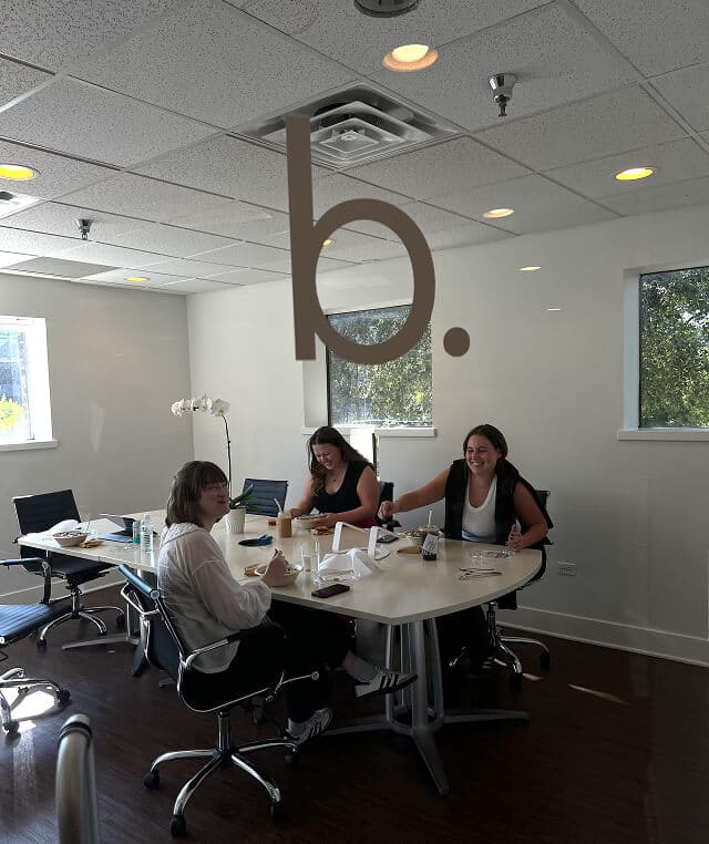 Three women sitting around a white conference table in an office, enjoying food and drinks, with windows and an orchid plant in the background.