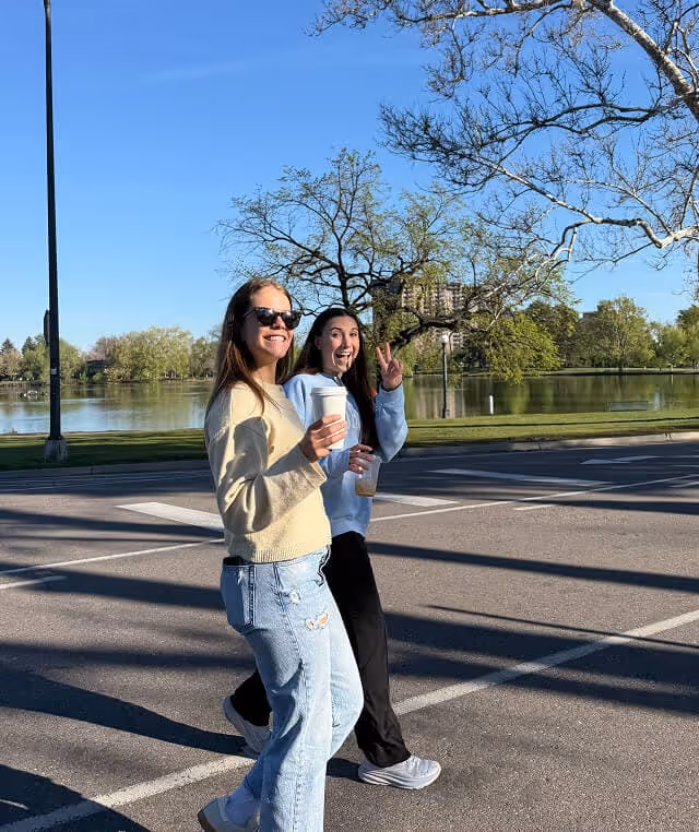 Two young women walking outdoors near a lake, one holding a coffee cup and the other making a peace sign, both smiling.