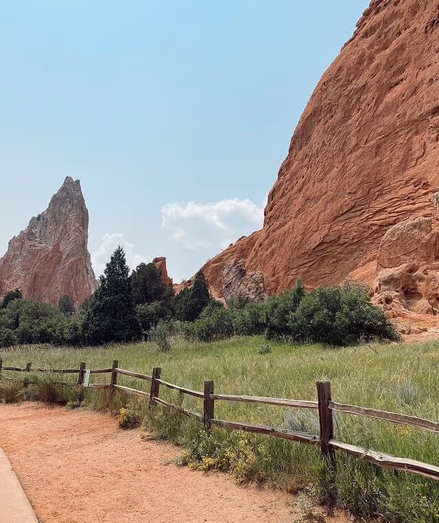 Dirt path bordered by a wooden fence with green bushes and large red rock formations under a clear blue sky.