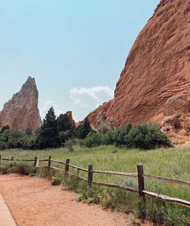 Dirt path bordered by a wooden fence with green bushes and large red rock formations under a clear blue sky.
