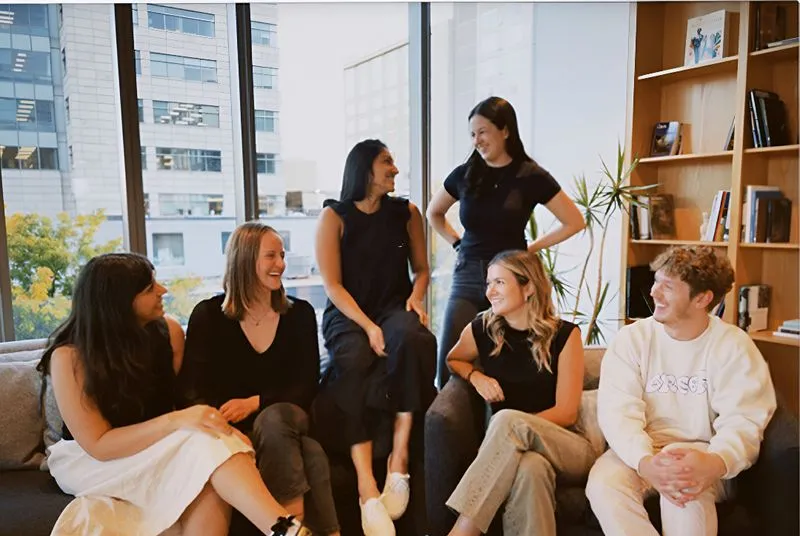 Group of six young adults sitting and standing in a modern office with large windows and bookshelves, engaging in a lively conversation.