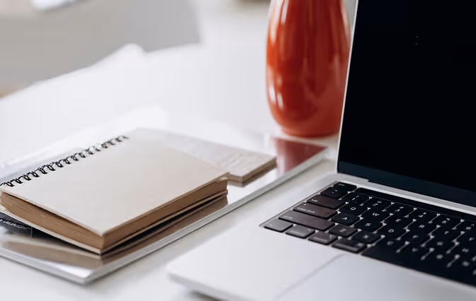 Close-up of a laptop keyboard and screen next to a stack of closed notebooks and a red vase on a white desk.