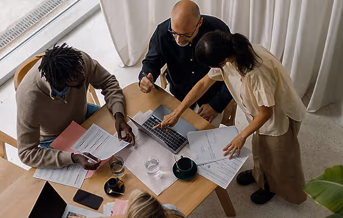 Three people gathered around a table reviewing documents and using a laptop during a meeting.