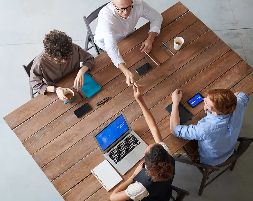 Four colleagues sitting around a wooden table in a meeting, with one woman reaching up to shake hands with a man standing beside the table.