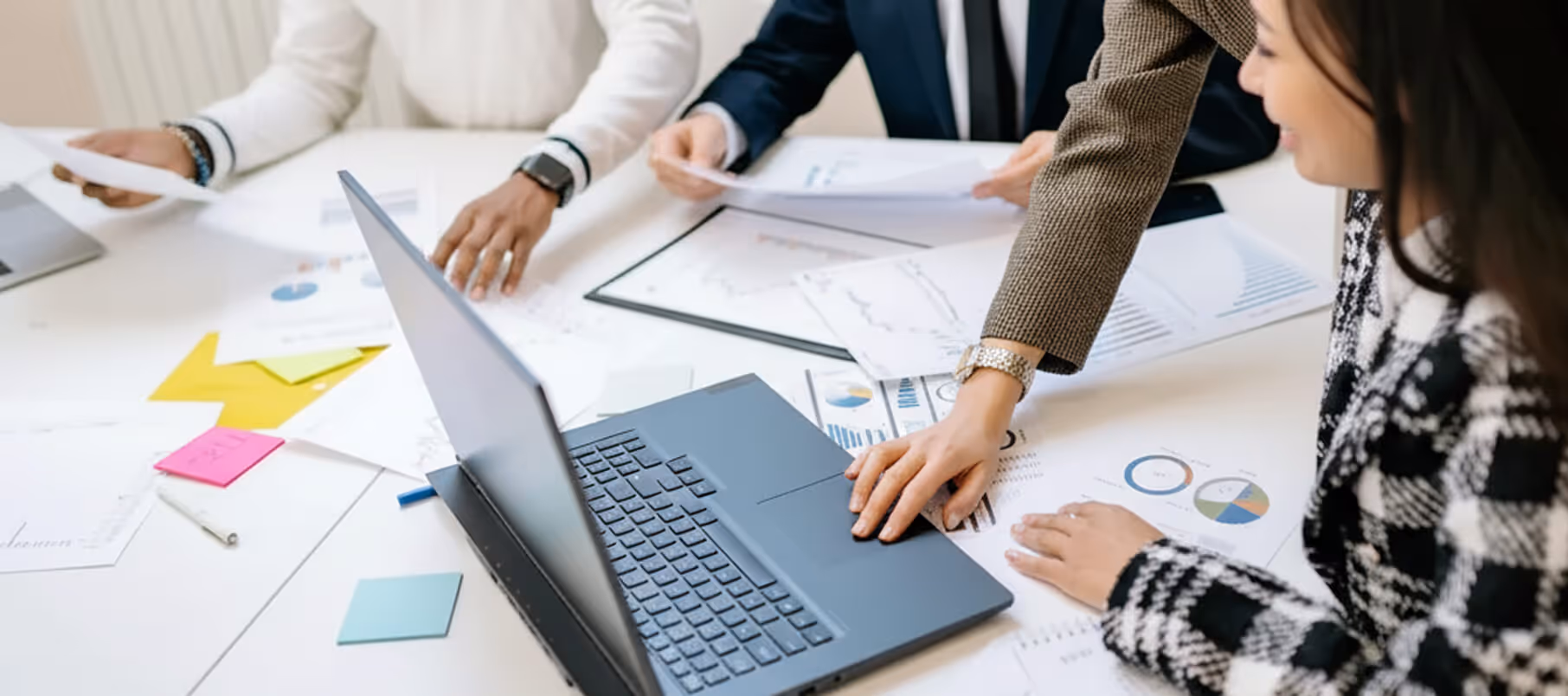 Three people collaborating at a table with a laptop and documents featuring charts and graphs.