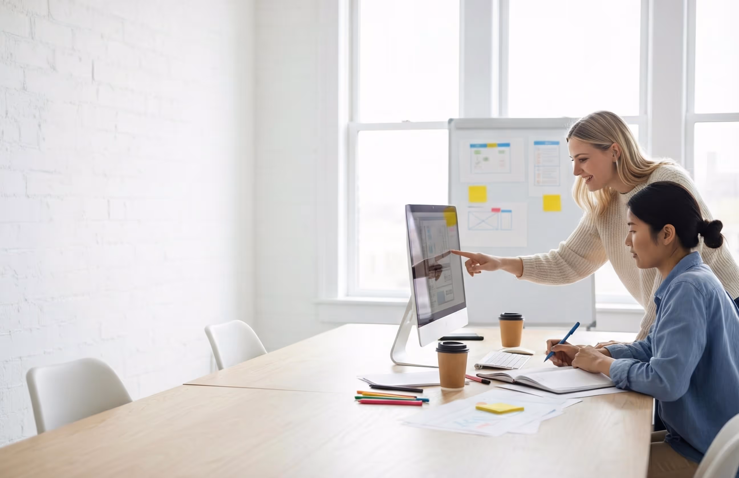 Two women collaborating at a desk in a bright office, one pointing at a computer screen while the other takes notes.