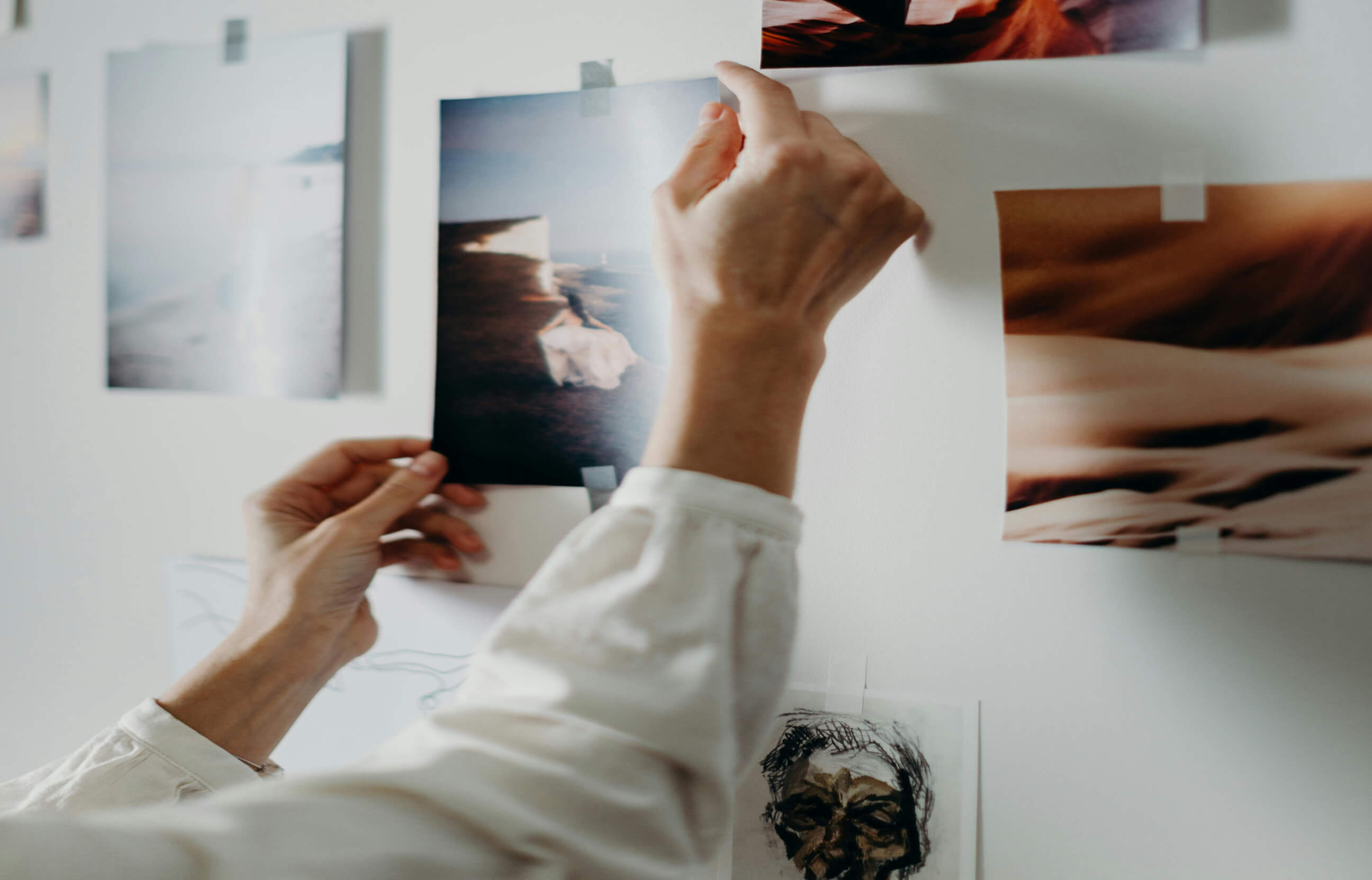 Person's hands taping a photo of a person on a coastline to a white wall with other photos and drawings.