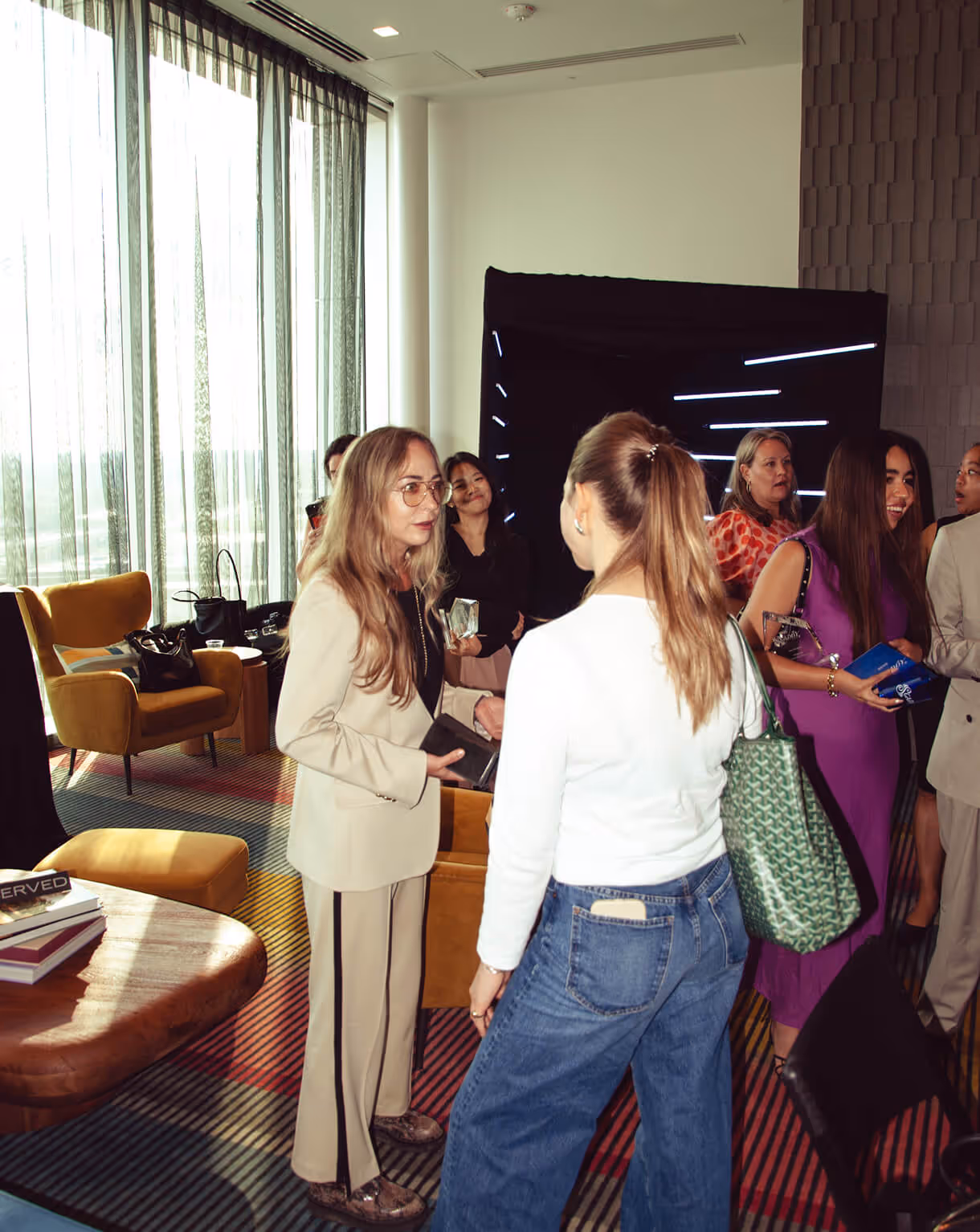 Women networking indoors with large windows and yellow chairs in background.