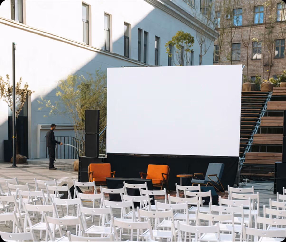 Empty outdoor event space with white chairs, stage with large blank screen, and a person watering plants.